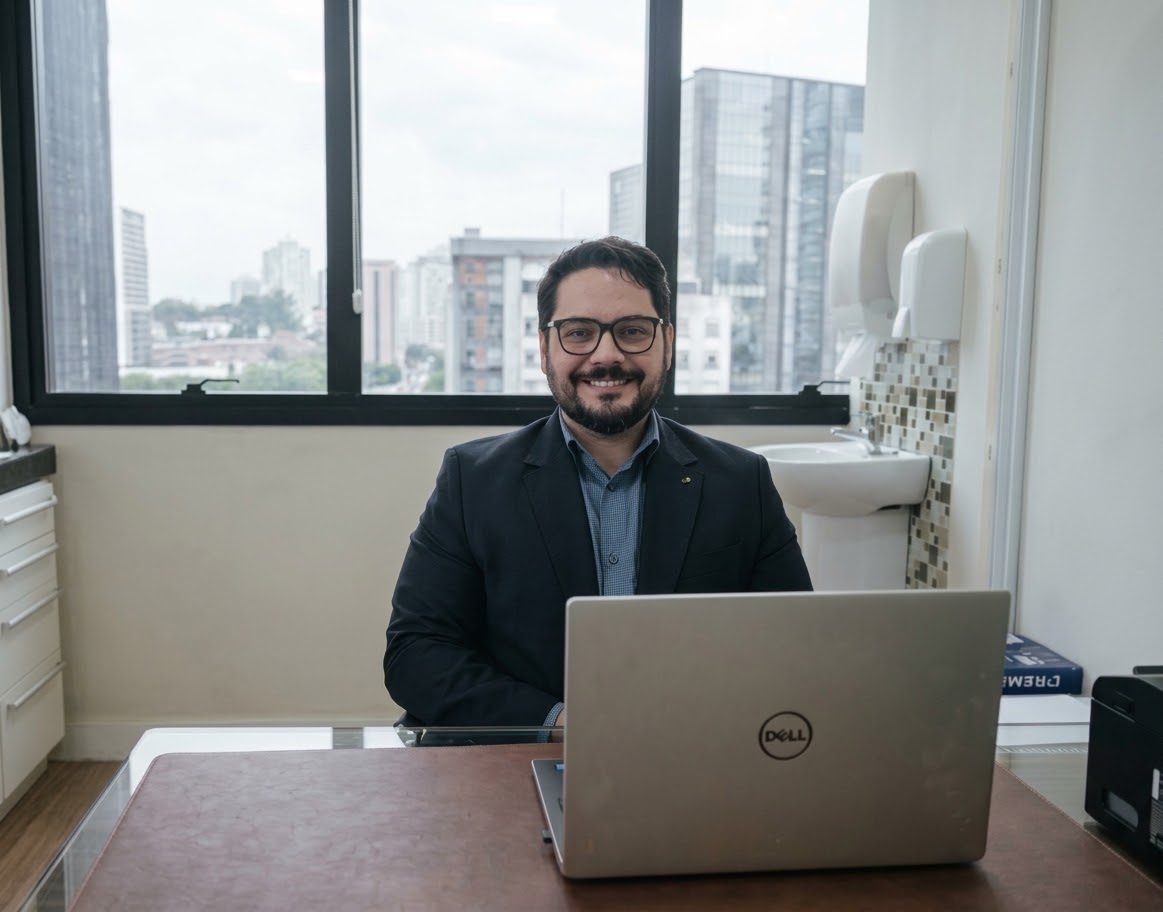 Dr. Daniel Caldeira Thomé em seu consultório na Poliklinik, sentado à mesa, com a janela e os prédios de São Paulo ao fundo